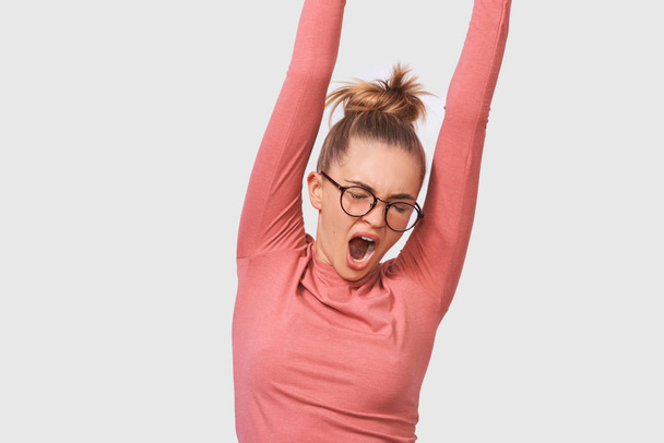 Image of tired Caucasian young woman yawning, wearing pink blouse and round transparent eyewear, posing over white studio wall. Sleepy female with hair bun, feel sleepy. People and emotions concept - Photo, image