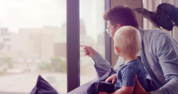 Young millennial father sitting on couch with baby daughter laughing and looking out apartment window in city  - Filmagem, Vídeo