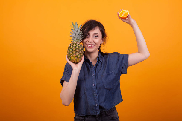 黄色の背景の上にスタジオでトロピカルフルーツを持つ若い美しい女性を笑う - 写真・画像