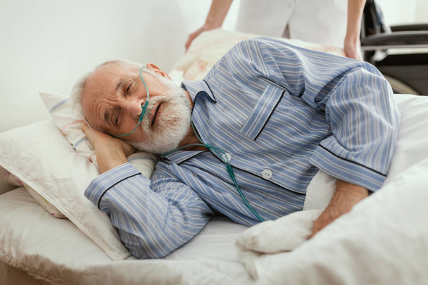 Sick elderly man wearing blue pajama lying in bed at nursing home - 写真・画像