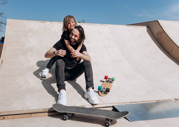 Happy young father and his son dressed in the stylish casual clothes are sitting in an embrace together on the slide next to the skateboards in a skate park at the sunny warm day - 写真・画像