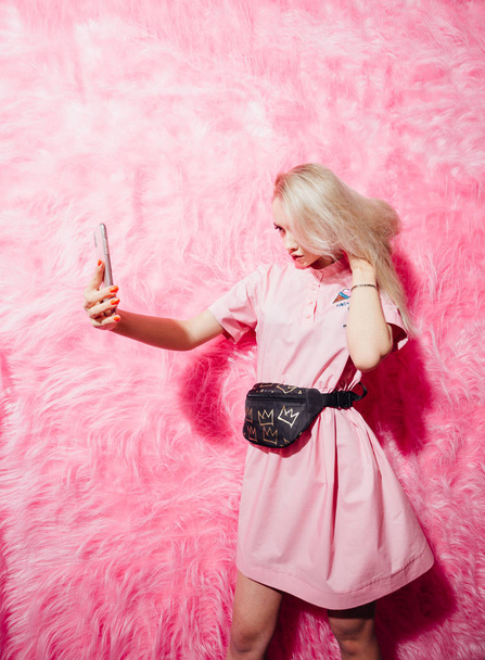 Beautiful young girl blogger dressed in fashion pink dress make a selfie on the background of pink fur wall in the show room - Фото, изображение