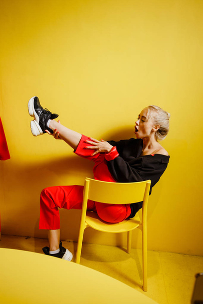 Young fashion girl blogger dressed in red trousers and black jacke possing sitting on the stool in the room with yellow walls and furniture - Foto, Imagem