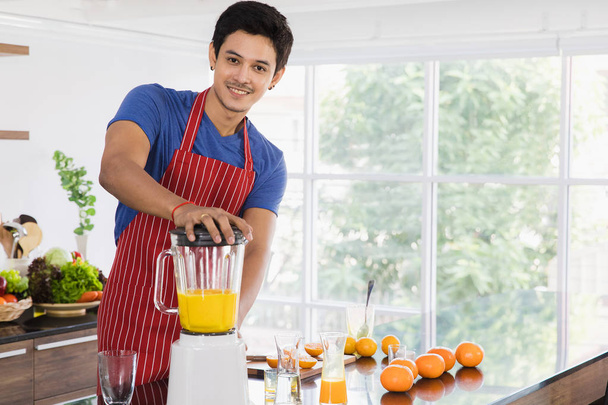 Front view shot of happy young Asian handsome bearded man in blue t shirt and striped apron use vegetable blender preparing healthy fresh orange juice for veggie meals in modern kitchen background - Foto, afbeelding