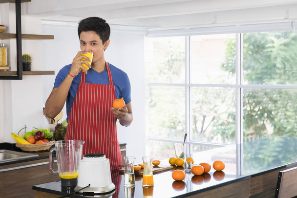 Első nézet lövés boldog fiatal ázsiai jóképű szakállas férfi kék pólóban és csíkos kötény áll a konyhában, és iszik frissen facsart narancslé, minden idők legjobb egészséges vegetáriánus ételeket - Fotó, kép