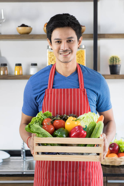 Close up shot of happy young Asian handsome bearded man wearing blue t shirt and striped apron hold a plate full of vegetables, presenting healthy meals images standing with modern kitchen background - Fotó, kép