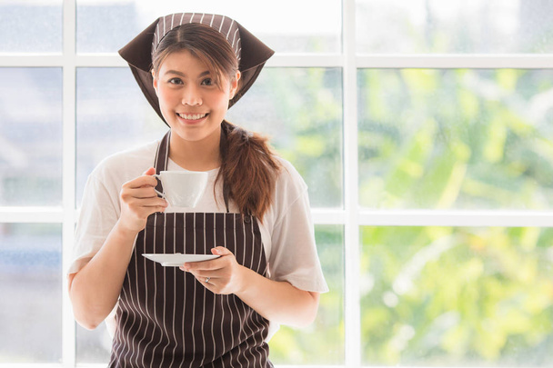 Çizgili yemek şapkası ve önlüğü takmış mutlu, genç ve güzel Asyalı kadın portresi modern mutfak arka planında kahve molası veriyor, büyük cam pencereli, kameraya gülümsüyor. - Fotoğraf, Görsel