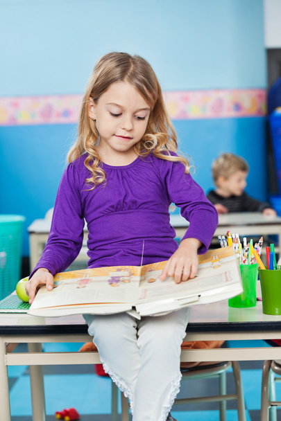 Girl Reading Book While Sitting On Desk At Kindergarten - Zdjęcie, obraz