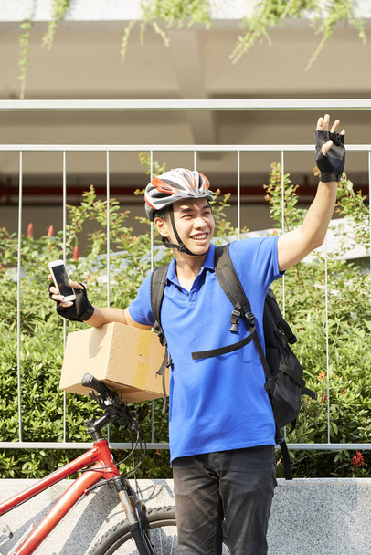 Portret van vrolijke koerier staande in de buurt van zijn fiets met kartonnen doos op stoel - Foto, afbeelding
