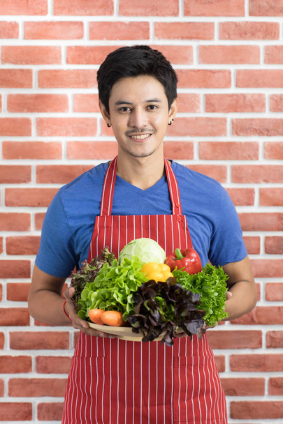 Ritratto di felice giovane asiatico bell'uomo barbuto indossando t shirt blu e grembiule a righe tenere un piatto pieno di verdure, presentando pasti sani immagini in piedi contro sfondo muro di mattoni rossi
 - Foto, immagini