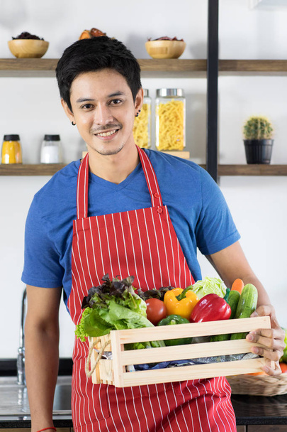 Detailní záběr šťastný mladý asijský pohledný vousatý muž na sobě modré tričko a pruhované zástěry držet talíř plný zeleniny, představovat zdravé jídlo obrázky stojící s moderním kuchyňským pozadím - Fotografie, Obrázek