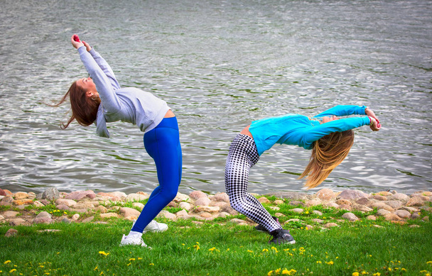 Δύο Καυκάσιες γυναίκες σε καλσόν και αθλητικά παπούτσια κάνει ασκήσεις γιόγκα στο φόντο του ποταμού. Εξάσκηση και δημιουργία μιας επιδεικικής σύνθεσης, ενώ στέκεται στην πόζα της γέφυρας ενός αθλητή.  - Φωτογραφία, εικόνα
