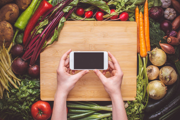 Hands with Smartphone over Fresh Organic Vegetables Background. Vegetarian Food Recipes. - Фото, изображение