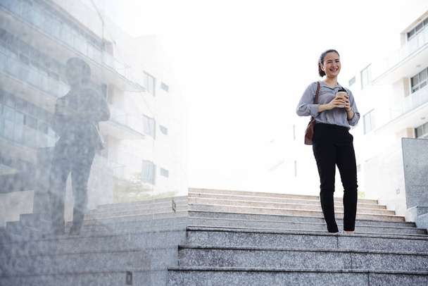 Happy young Asian woman with cup of coffee walking down the stairs  - Photo, Image