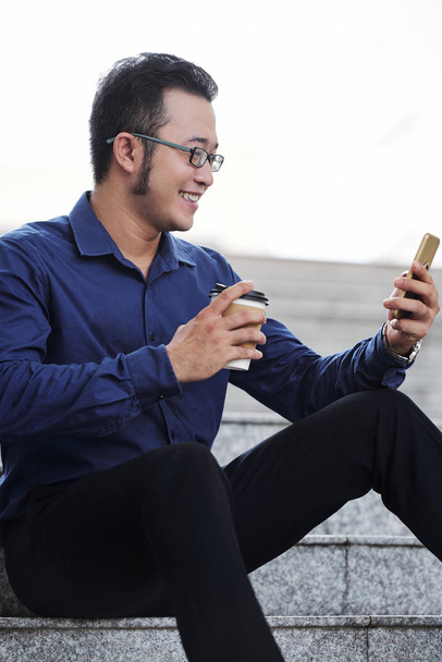 schöner junger vietnamesischer Geschäftsmann sitzt auf der Treppe, trinkt Kaffee und liest SMS  - Foto, Bild