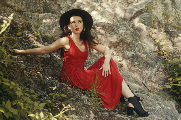 ファッショナブルな女の子が岩の上に座っています。ファッショナブルな赤いロングドレスを持つヒップスターの女性。山の中の少女美しい黒い帽子をかぶったブルネット。赤い唇と美しいイヤリングを持つ女の子 - 写真・画像