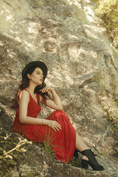 Muodikas tyttö istuu jäillä. Hipster nainen muodikas punainen pitkä mekko. Tyttö vuorilla. Ruskeaverikkö kauniissa mustassa hatussa. Tyttö punaiset huulet ja kauniit korvakorut
 - Valokuva, kuva
