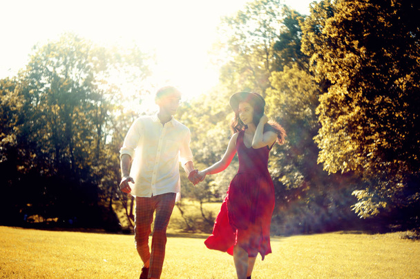 Couple courant dans la forêt, champ. Fille dans une longue robe rouge et chapeau, homme élégamment habillé. Coucher de soleil dans le parc. La fille et le gars marchent. Rendez-vous romantique parfait. Un homme en chemise blanche en été
. - Photo, image