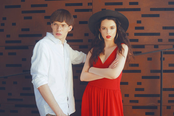 Beautiful fashionable couple on an abstract background. Fashionable hipsters. Girl in a red dress and black big hat. A man with glasses and a white shirt. Model pouse. - Photo, Image