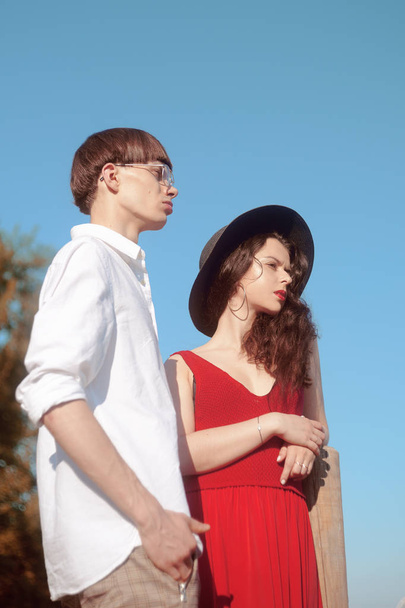 Beautiful fashionable couple on the beach near the river. Fashionable hipsters. Girl in a red dress and black big hat. A man with glasses and a white shirt. Model pouse. Enjoy a sunny summer day. - Photo, Image