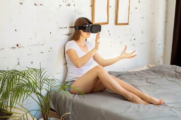 hermosa mujer joven en una camiseta blanca se sienta en la cama con gafas de realidad virtual en el interior
 - Foto, Imagen