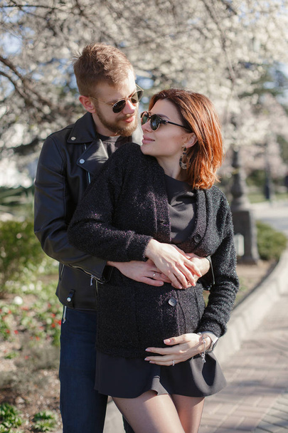 A couple in love, a man and a pregnant woman, walk together in the spring in a beautiful sunny city.Pregnant woman and man hug each other and show their love. Happy couple is expecting a baby. The concept of love and family well-being - Fotografie, Obrázek