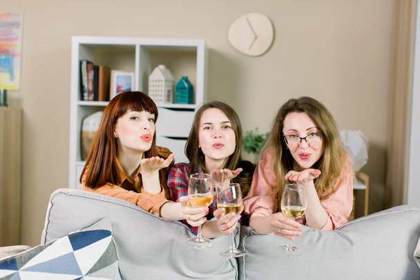 Three happy women blowing air kiss and holding glasses with wine, sitting on the floor behind gray sofa in cozy stylish room at home - Fotografie, Obrázek