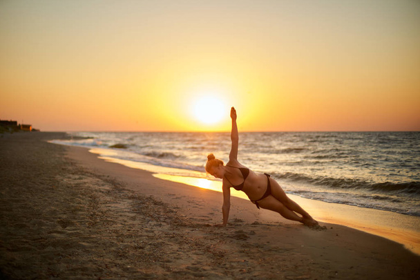 Mujer auténtica en traje de baño haciendo yoga vasisthasana en la playa por la mañana. Silueta de chica de forma real sin retocar en la tabla lateral asana cerca del mar al amanecer. Entrenamiento femenino positivo en el atardecer
 - Foto, imagen