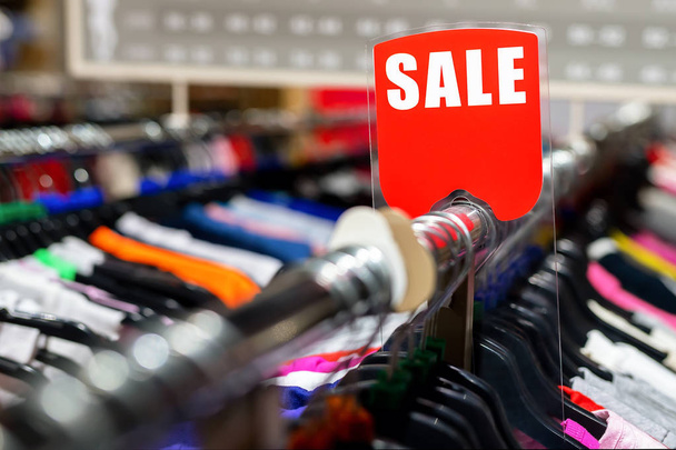 Retail clothes store clearance. Garment shop with various bright youth casual wear at discount price. Wear hangers and red SALE signboard. Seasonal sales on fashion market - 写真・画像