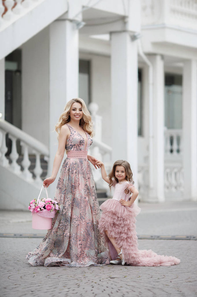 Beautiful slender woman and her little daughter,having fun together outdoors in the spring,on a palace alley with flowering peaches,dressed in beautiful ball gowns of pink color.Mom and daughter in beautiful evening dresses on the street - Фото, изображение