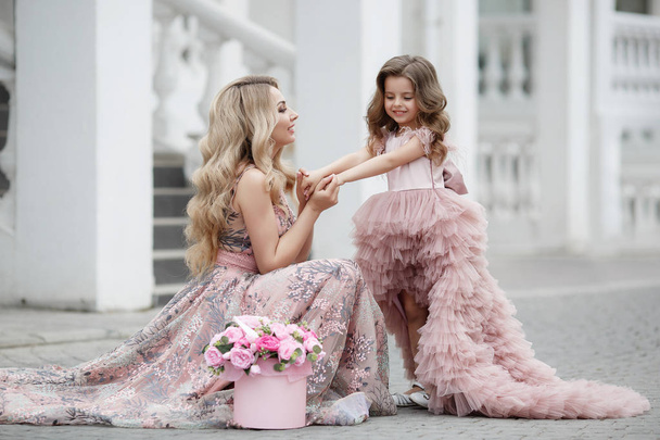 Beautiful slender woman and her little daughter,having fun together outdoors in the spring,on a palace alley with flowering peaches,dressed in beautiful ball gowns of pink color.Mom and daughter in beautiful evening dresses on the street - Photo, image