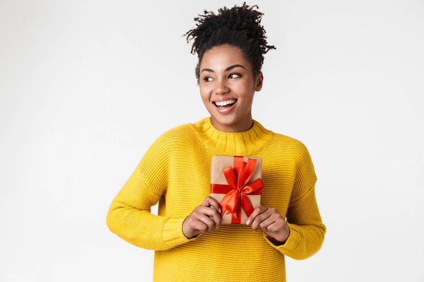 美しい若いアフリカの興奮感情的な幸せな女性は、プレゼントボックスを保持する白い壁の背景の上に孤立したポーズ. - 写真・画像