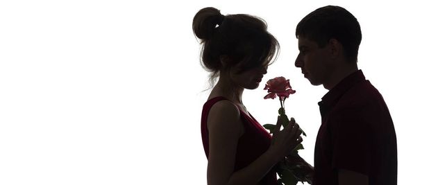 silhouette profile of a young couple in love on white isolated background, man giving a woman a rose flower, concept love and romance - Фото, изображение
