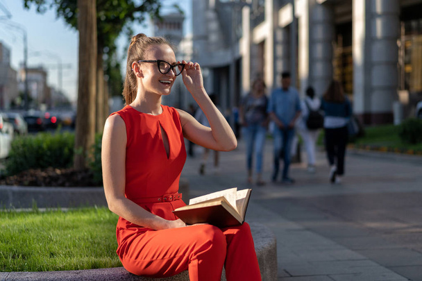 a young stylish european girl in a red suit and glasses sits on the parapet on the street and reads a book. Past her people pass, you can see the city life. There is a method for a small signature. - Foto, immagini