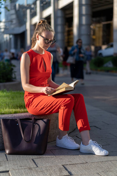 a young stylish european girl in a red suit and glasses sits on the parapet on the street and reads a book. Past her people pass, you can see the city life. There is a method for a small signature. - Fotó, kép