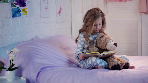 słodkie dziecko preteen w piżamie siedzi na łóżku z misia, czytanie książki, uśmiechając się i wyraża zdumienie - Materiał filmowy, wideo