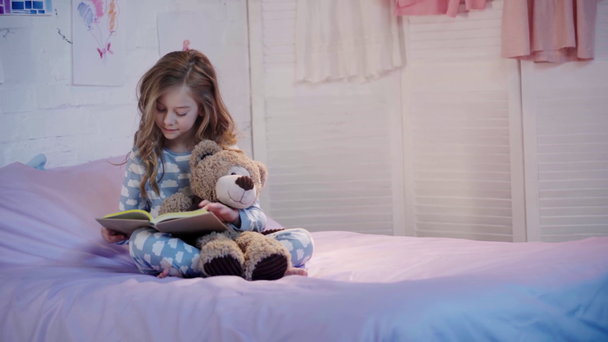 aranyos preteen gyermek pizsama ül az ágyon a mackó és nevetve olvasás közben könyv - Felvétel, videó