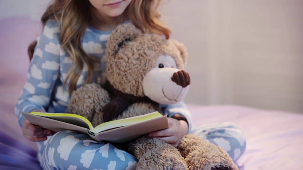 vista cortada de criança pré-adolescente em pijama sentado na cama com ursinho de pelúcia e livro de leitura
 - Filmagem, Vídeo