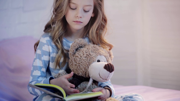 cute preteen child in pajamas sitting on bed with teddy bear and reading book - Footage, Video