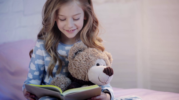 schattig Preteen kind in pyjama zittend op bed met teddybeer en lachen tijdens het lezen van boek - Video