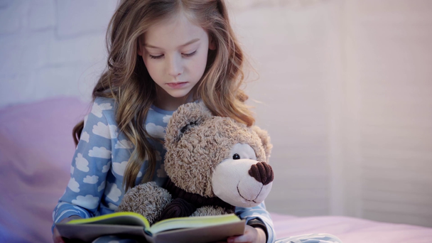 lindo niño preadolescente en pijama sentado en la cama con oso de peluche, sonriendo y expresando asombro al leer el libro
 - Metraje, vídeo