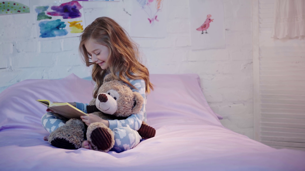 χαριτωμένο Προεφηβικό παιδί με πιτζάμες που κάθονται στο κρεβάτι με αρκουδάκι και χαμογελώντας ενώ διαβάζοντας το βιβλίο - Πλάνα, βίντεο