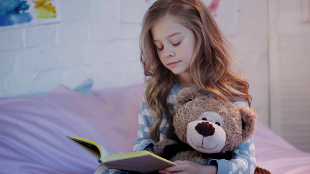 roztomilá dětská mládež v pyžamu, která sedí na posteli s medvídkem a usmívá se při čtení knihy - Záběry, video