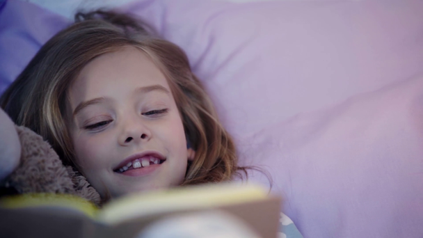 χαριτωμένο Προεφηβικό παιδί με πιτζάμες που κείτεται στο κρεβάτι με αρκουδάκι και χαμογελώντας ενώ διαβάζοντας το βιβλίο - Πλάνα, βίντεο