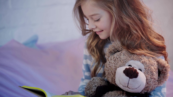 aranyos preteen gyermek pizsama ül az ágyon a mackó és nevetve olvasás közben könyv - Felvétel, videó