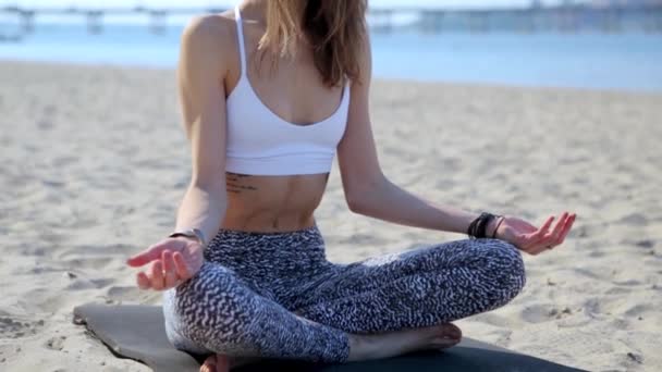 Portrait of a beautiful relaxed young woman sitting in lotus pose at the beach with sea and city background. womans hands in namaste murda pose - Footage, Video