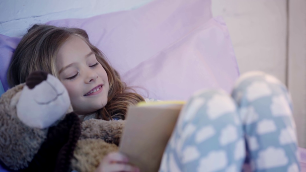 cute preteen child in pajamas lying on bed with teddy bear and smiling while reading book - Footage, Video