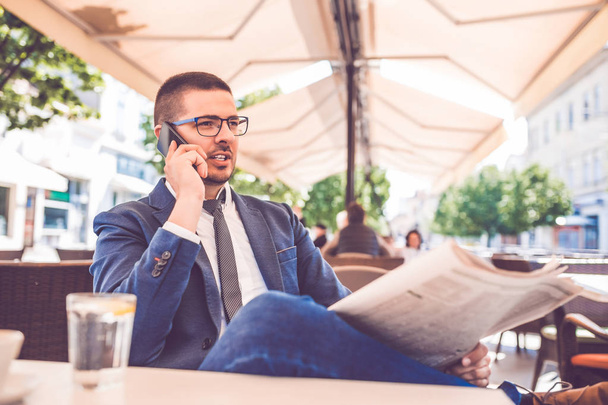 Nuori liikemies lukee sanomalehtiä ja puhuu matkapuhelimella
 - Valokuva, kuva