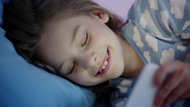 cheerful kid in pajama lying in bed and using smartphone with smile - Záběry, video