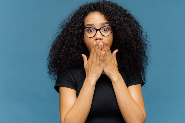 Photo of stupefied young female covers mouth, has bated breath, bugged eyes, Afro hair, cannot believe in shocking news, wears casual black t shirt, stands over blue background. Omg concept. - Photo, Image